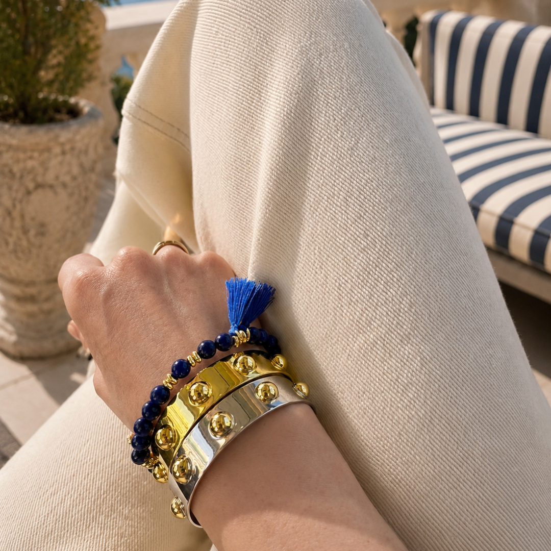 Silver and Gold Roman cuffs layered with a lapis lazuli breaded bracelet with blue tassels on neutral Riviera styling background