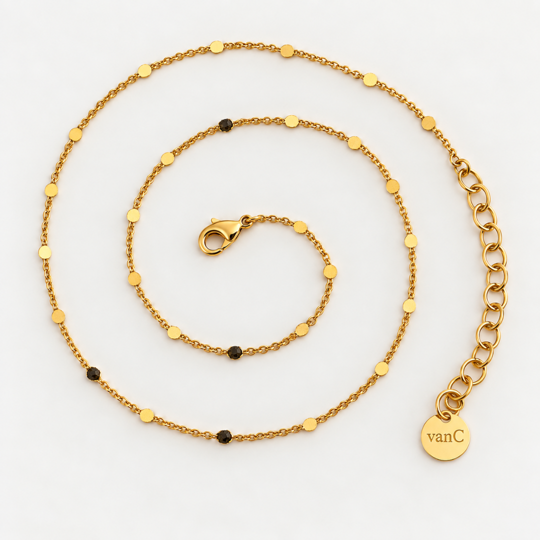 Gold plated Dotty chain from
Blauw van C with small flat gold discs, displayed in a spiral on a white background. A round brand tag engraved with 'vanC' logo is attached to the clasp.