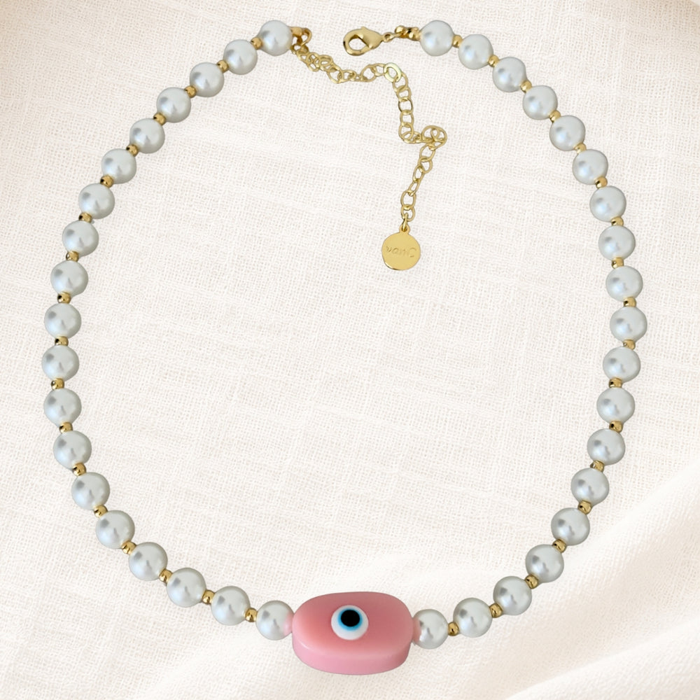 Gold plated pearl necklace with soft pink evil eye pendant on white linen background.