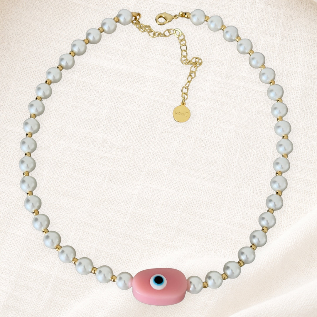 Gold plated pearl necklace with soft pink evil eye pendant on white linen background.