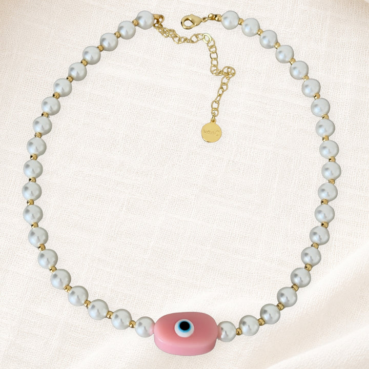 Gold plated pearl necklace with soft pink evil eye pendant on white linen background.