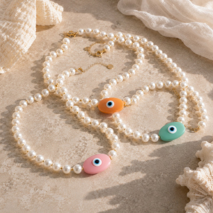 Three 18K Gold plated pearl necklaces with colourful handcrafted evil eye bead charms in pink, peach and aqua on stone and linen background.