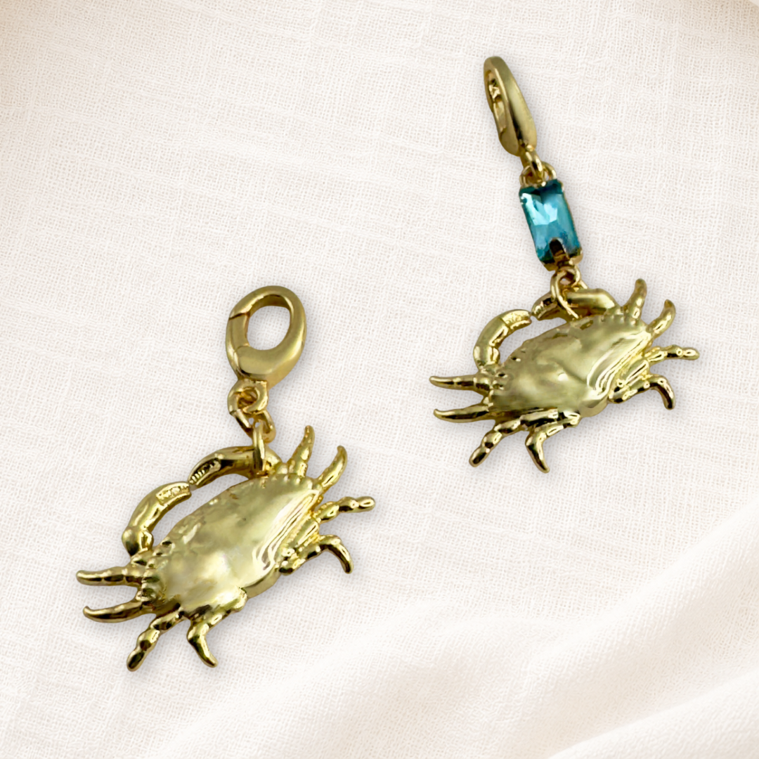 Two gold plated lucky crab charms with lobster clasps. One plain gold and the other with blue crystal, displayed on a cream textured fabric background.