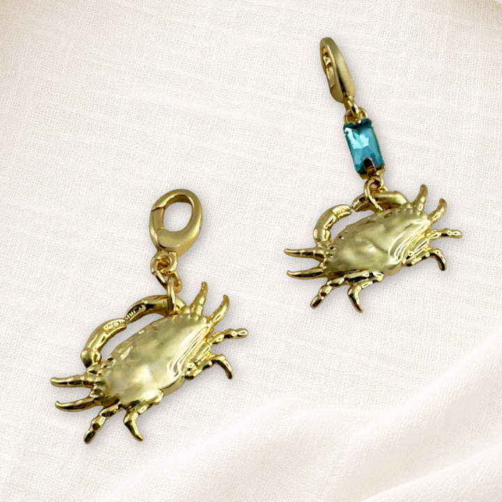 Two gold plated lucky crab charms with lobster clasps. One plain gold and the other with blue crystal, displayed on a cream textured fabric background.