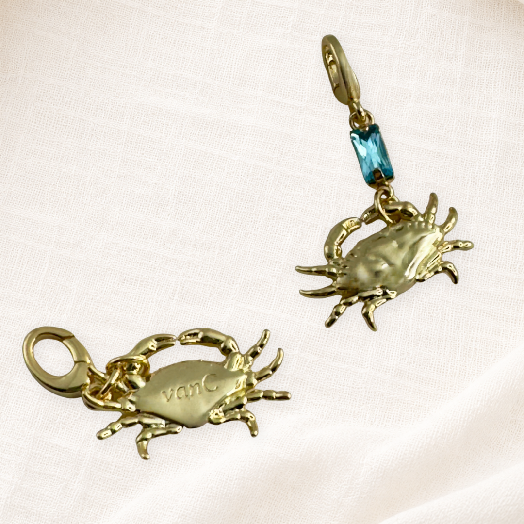 Two gold plated lucky crab charms with lobster clasps. One plain gold with brand logo and the other with blue crystal, displayed on a cream textured fabric background.
