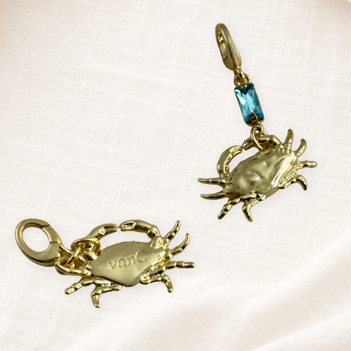 Two gold plated lucky crab charms with lobster clasps. One plain gold with brand logo and the other with blue crystal, displayed on a cream textured fabric background.