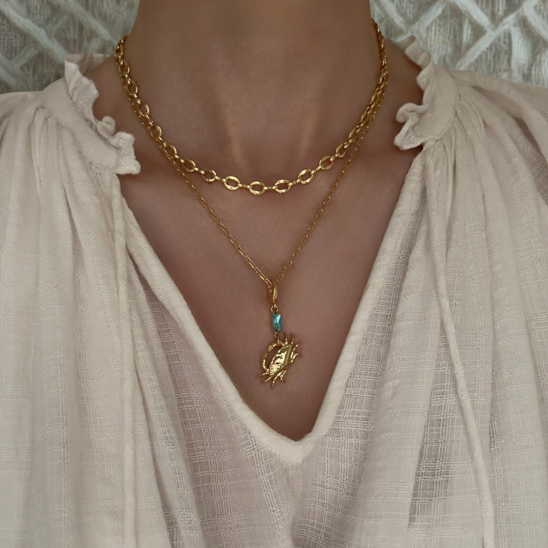 A close-up of a person wearing layered gold necklaces, including a short boho chain, a longer chain with the Lucky Crab pendant with blue crystal paired with an off white linen bohemian blouse.