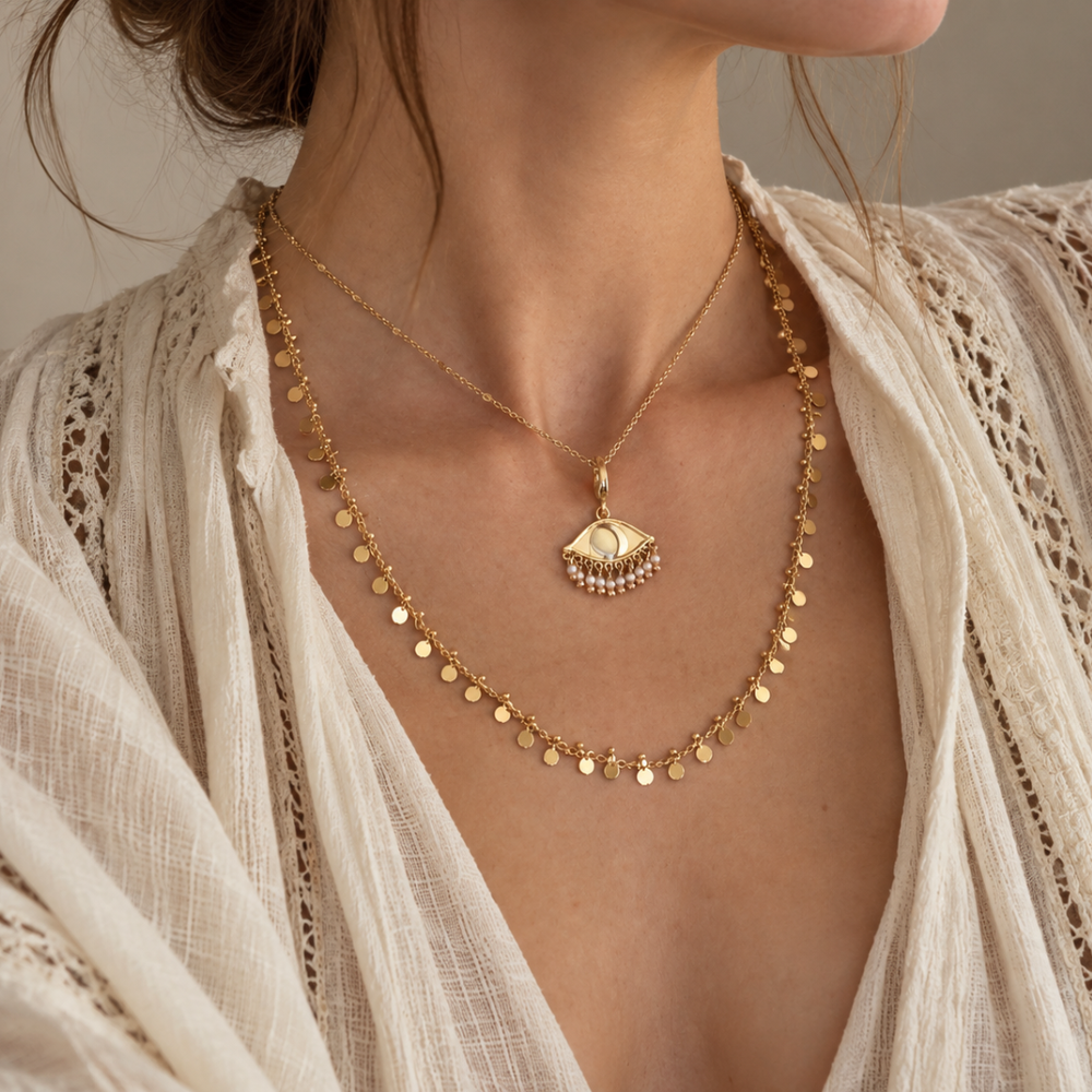 A close-up of a model wearing a layered gold plated chains look from Blauw van C. The top layer features the Dotty chain with See the world small gold pendant with pearls. The bottom layer is the Magic chain adorned with small dangling gold coins (discs). 