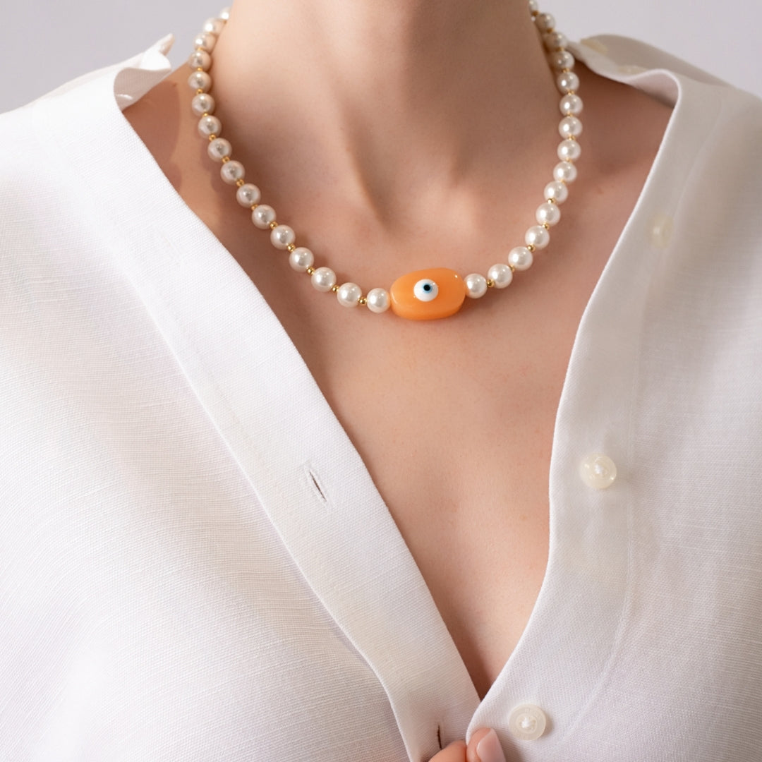 Model wearing 18K Gold plated pearls necklace with central evil eye pendant in peach with a white linen shirt.