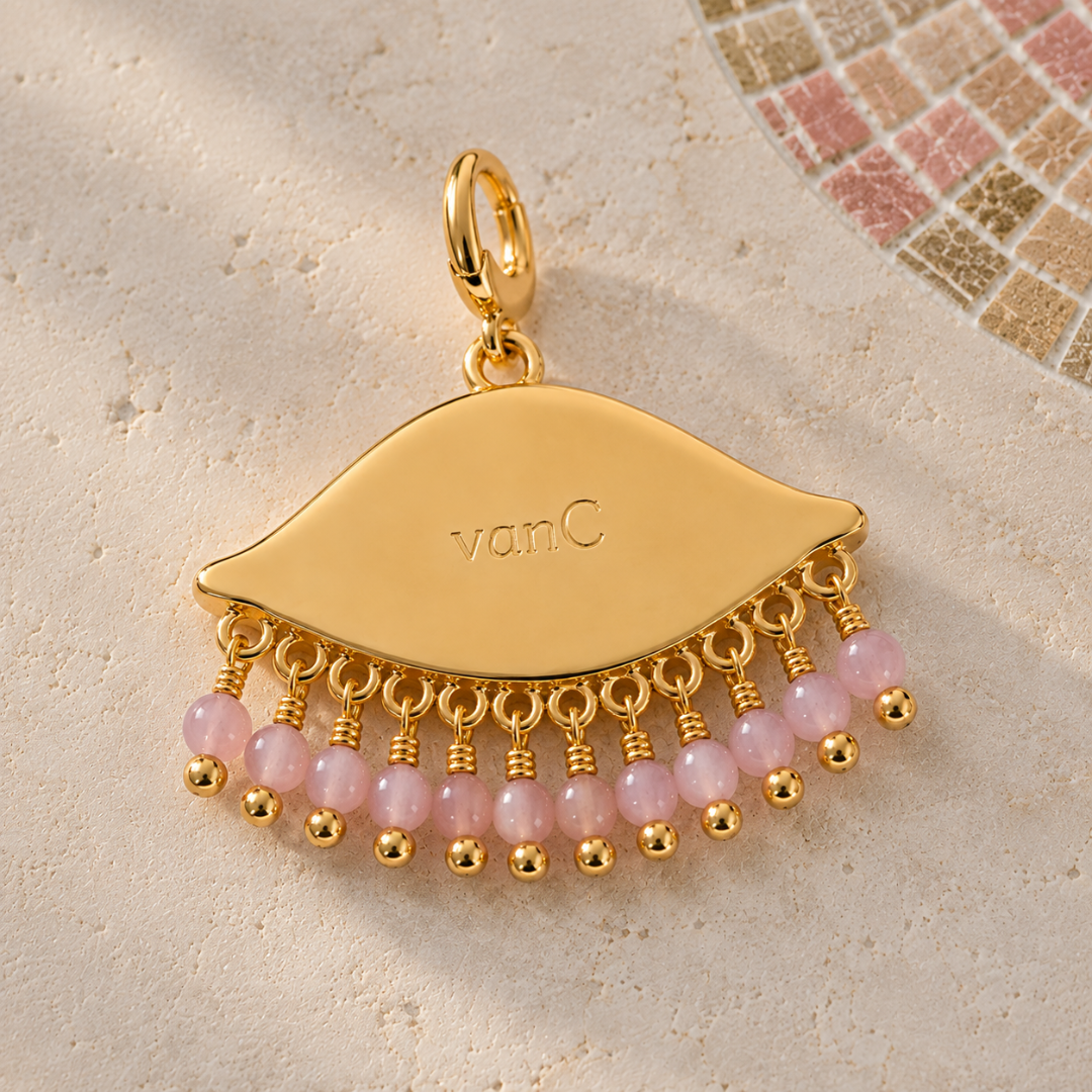 See the world gold-plated eye-shaped pendant charm with dangling pink beads, photographed on a light textured surface with a small mosaic tile detail in the upper right. The pendant is engraved with the brand logo 'vanC'.
