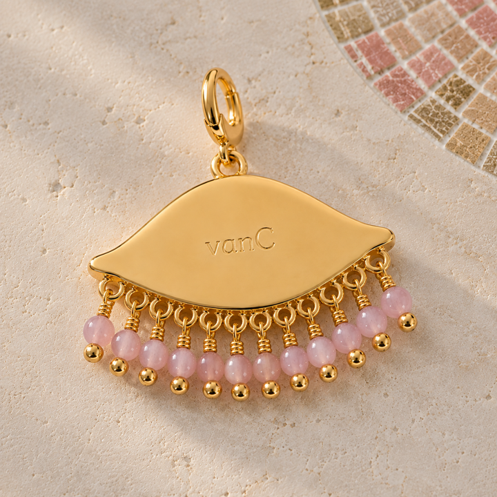 See the world gold-plated eye-shaped pendant charm with dangling pink beads, photographed on a light textured surface with a small mosaic tile detail in the upper right. The pendant is engraved with the brand logo 'vanC'.