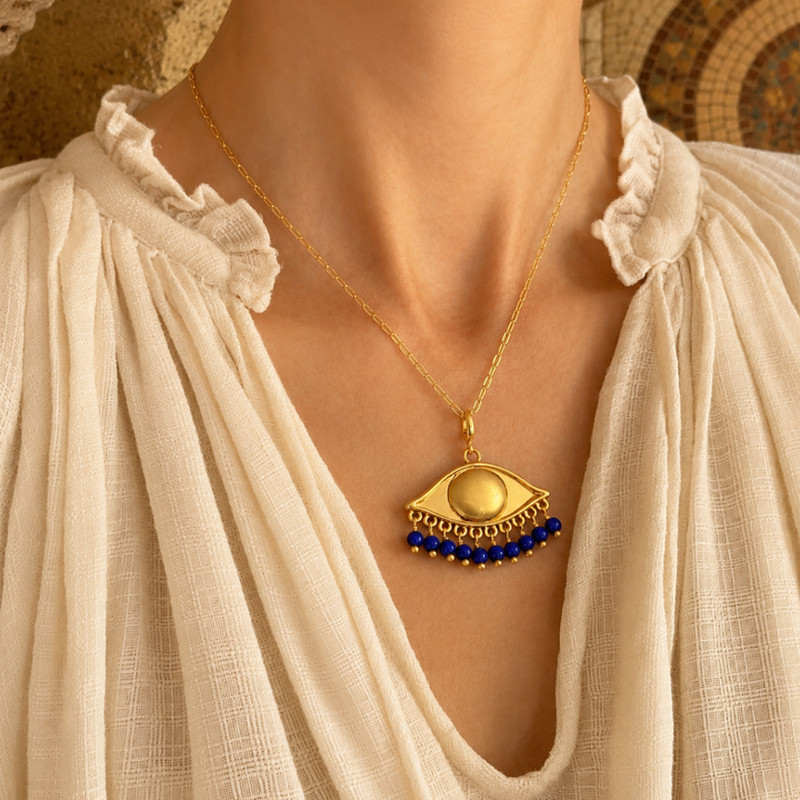A gold-plated eye-shaped pendant necklace with cobalt beads, worn on a model's neck with a bohemian linen top in a warm ancient light.