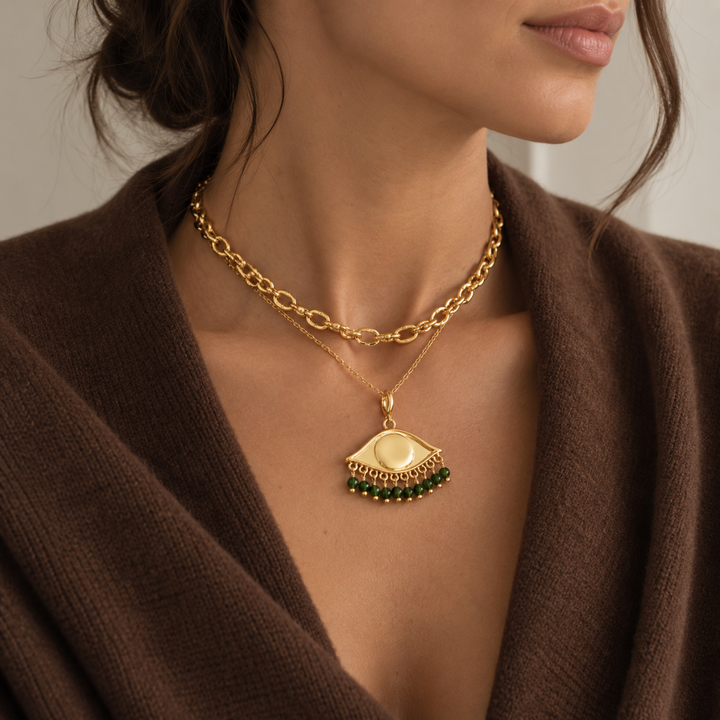 A close-up of a model wearing a layered gold necklace look, featuring a Boho gold plated chain and a delicate chain with See the world  eye charm  with green bead fringe. Styled with a brown cashmere top. 