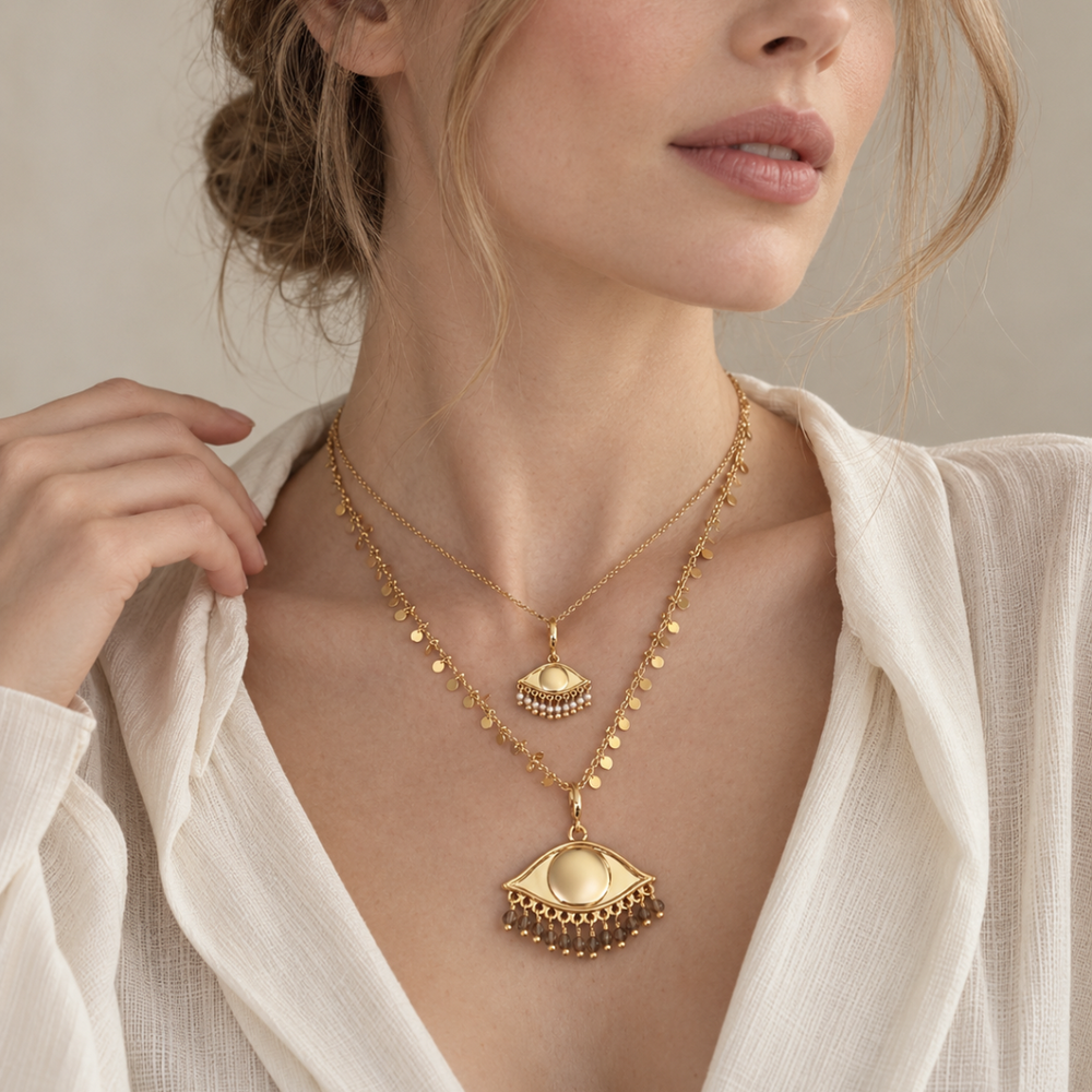 A close-up of a woman wearing layered gold plated chains , styled with See the world charms pendants in two sizes and an off white top.