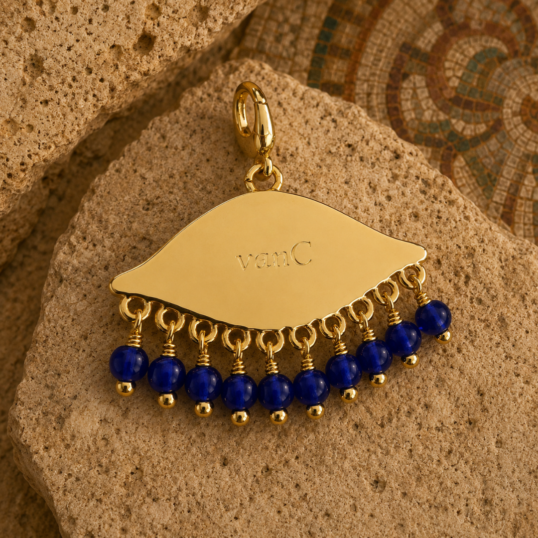 Back view of the See the World gold-plated eye pendant with cobalt blue beads, engraved with the vanC logo, photographed on warm natural stone with an ancient mosaic background.