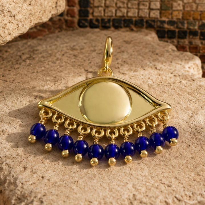 Gold plated oversized eye pendant charm with dangling cobalt blue beads, displayed on an old stone surface with a mosaic background. 