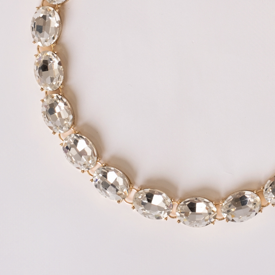 Close up detail of gold-plated necklace with white crystals on a light gray background.