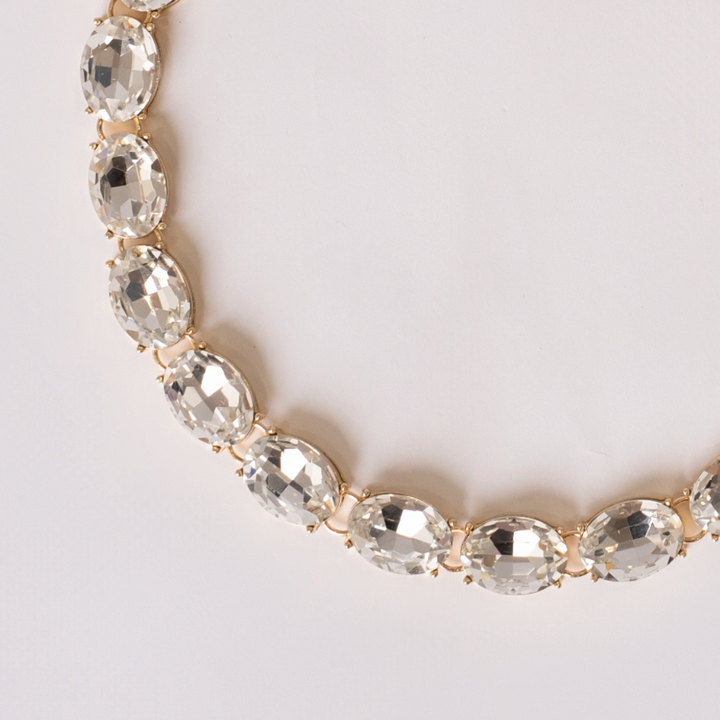 Close up detail of gold-plated necklace with white crystals on a light gray background.