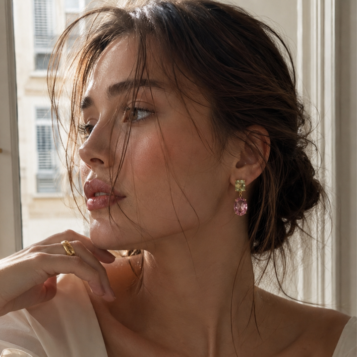 A close-up lifestyle shot of a European model with dark hair in a low bun, wearing gold plated light pink and green crystal drop earrings. Editorial shot, the model is looking to the side in a Paris flat. 