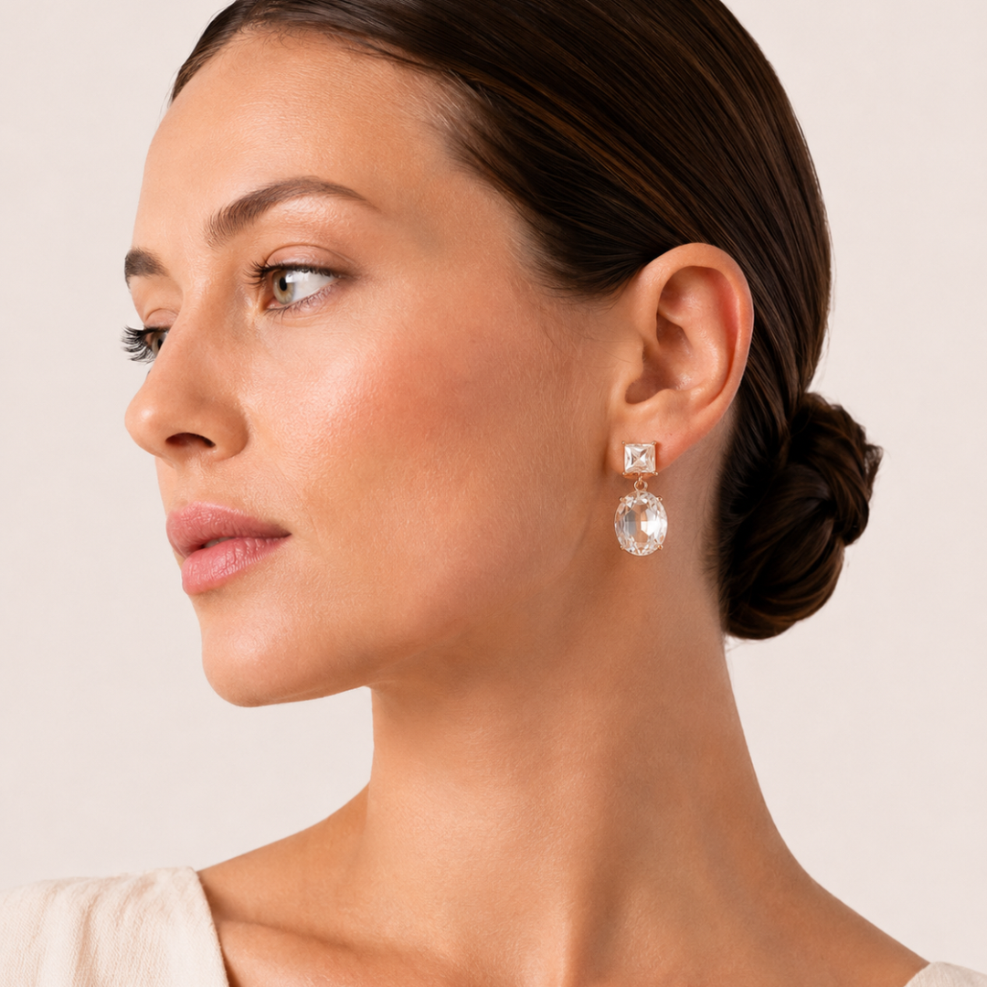 A close up profile of a woman wearing Two Way Glow clear crystal drop earrings with square and oval crystals in 18K gold-plated settings.