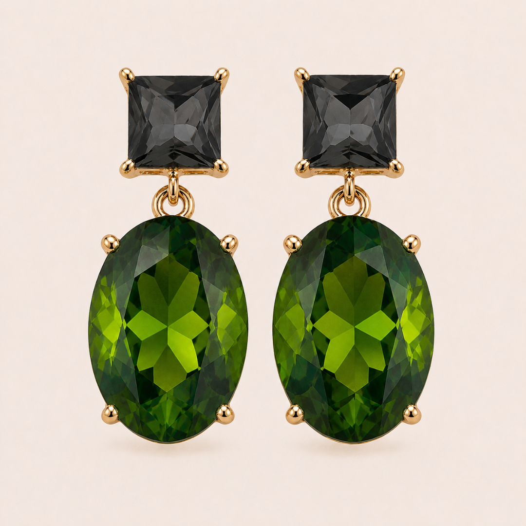 Close up photo of 18K gold plated  drop earrings featuring a square grey crystal at the top and an oval, faceted deep green oval crystal below, set against a solid pale neutral background.