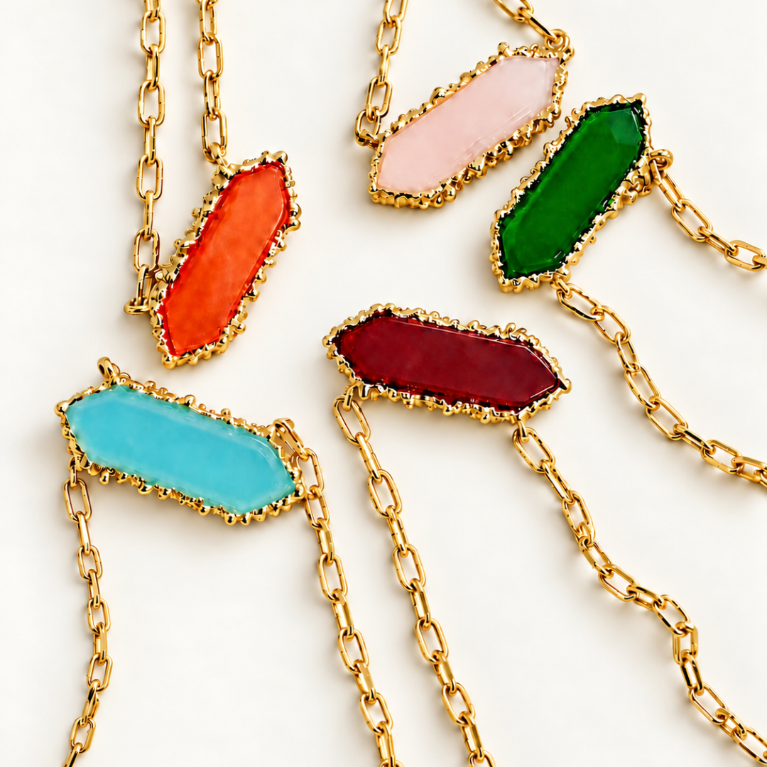 Close-up of five Blauw van C Andalus necklaces with orange, pale pink, green, red and blue agate pendants in decorative gold-plated settings on a plain off white background.